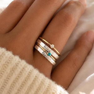 May include: A stack of three rings worn on a finger. The top ring is a thin gold band with a small clear stone. The middle silver band is engraved with the name Luca. The bottom silver band is engraved with the name Lexi and a blue stone.