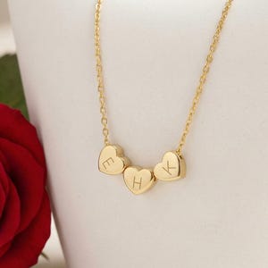 Può includere: Una collana dorata con tre ciondoli a forma di cuore, ognuno con una lettera. La collana è esposta su uno sfondo bianco, con una rosa rossa in primo piano. Un gioiello delicato.