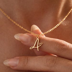 May include: A gold-toned necklace featuring a delicate chain and a pendant shaped like the cursive letter A, accented with a small heart symbol attached to the right side of the letter. The pendant is suspended from a thin, matching gold-toned chain.