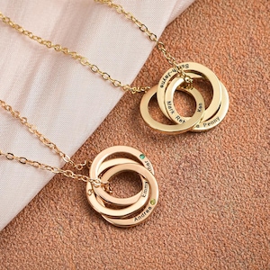May include: Gold-colored necklaces with interlocking rings. Each ring is engraved with a name. One necklace has a small green gem. The necklaces are displayed on a light pink cloth and a brown textured surface.