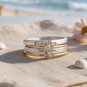 May include: A stack of two rings featuring a brushed silver band with the name Kinsley engraved and a smooth gold band with the name Peyton engraved. Each ring is accented with a small, round, light purple gemstone set in a gold bezel.