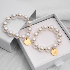 Può includere: Due braccialetti di perle con dettagli dorati e ciondoli con iniziali sono presentati in una scatola bianca. I braccialetti presentano grandi perle bianche rotonde e piccole perline dorate. Ogni braccialetto ha un ciondolo dorato con una lettera.