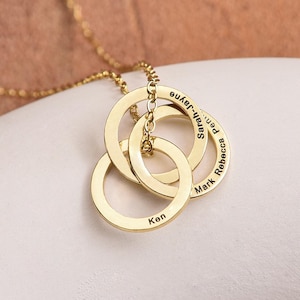 May include: Gold-colored necklace with three interlocking rings. Each ring is engraved with a name: Ken, Mark, Rebecca, Sarah-Jayne, and Pen. The necklace is displayed on a white surface.