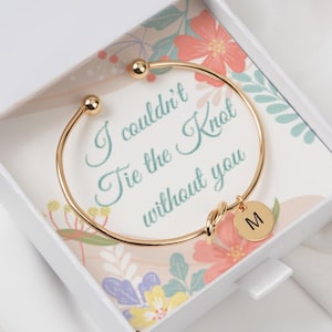 Puede incluir: Un brazalete dorado con un diseño de nudo y un dije redondo con la letra "M" se muestra en una caja de regalo blanca. La caja tiene un diseño floral y el texto "I couldn't Tie the Knot without you".