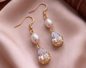 May include: A pair of gold-toned drop earrings featuring a white oval pearl suspended above a clear, pear-shaped faceted crystal. The crystal is encased in a delicate gold-toned filigree floral cap, and the earrings hang from gold-toned hooks.