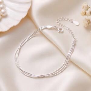 May include: A silver-toned multi-strand bracelet featuring three delicate snake chains. The bracelet includes a lobster claw clasp and an adjustable extender chain with a small teardrop-shaped charm at the end, creating a layered appearance.