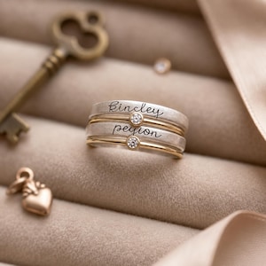 Può includere: Una pila di due anelli color argento con accenti color oro e pietre rotonde trasparenti. Ogni anello è inciso con un nome in corsivo nero, Kincley sulla fascia superiore e peyion su quella inferiore, con una piccola montatura dorata.