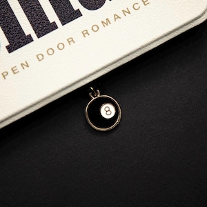 May include: A gold-toned charm featuring a black circle with a white number 8 inside. The charm is hanging from a black cord. The background is a white and black surface with the words "OPEN DOOR ROMANCE".
