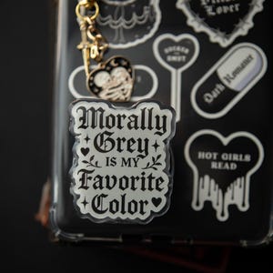 Może przedstawiać: Przezroczyste etui na telefon z różnymi naklejkami i złotym brelokiem. Naklejki zawierają serce z napisem "Villain Lover", pigułkę z napisem "Dark Romance" oraz naklejkę z napisem "Morally Grey is my Favorite Color".