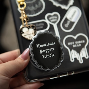 May include: Black phone case with stickers, including a pop socket that reads "Emotional Support Kindle" in white gothic font. The case has a gold charm and a heart-shaped charm. Other stickers include hearts and a pill.