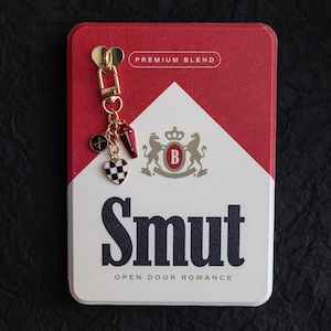 May include: A red and white rectangular case with the word "Smut" in large black letters. A gold-colored keychain with charms, including a checkered heart, hangs from the top. The text "Premium Blend" is above the case's white triangle.