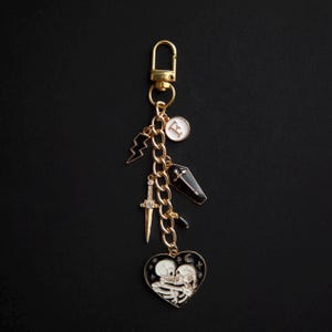 May include: A gold-colored keychain with multiple charms. Charms include a lightning bolt, a sword, a coffin, and a heart-shaped charm with two skeletons embracing. The keychain also has a charm with the letter "E".