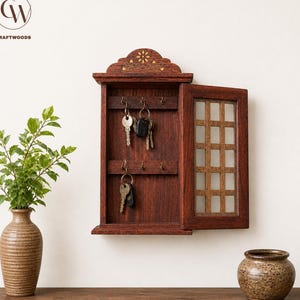 Puede incluir: Un armario para llaves de pared hecho de madera con acabado marrón oscuro, que presenta una puerta abatible con patrón de rejilla, ganchos metálicos internos para colgar llaves y una parte superior arqueada con detalles florales incrustados.