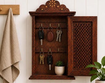 May include: A dark brown wooden wall-mounted key cabinet with a hinged door featuring a lattice pattern. The interior contains six brass hooks holding various keys and key fobs, along with a small white pot containing a green succulent plant.