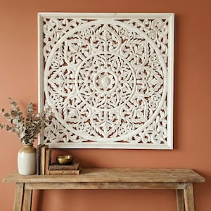 May include: A square white wooden wall panel featuring an intricate hand-carved floral mandala design with a central circular motif and symmetrical leaf patterns, framed by a simple border, mounted on a solid terracotta-colored wall surface.