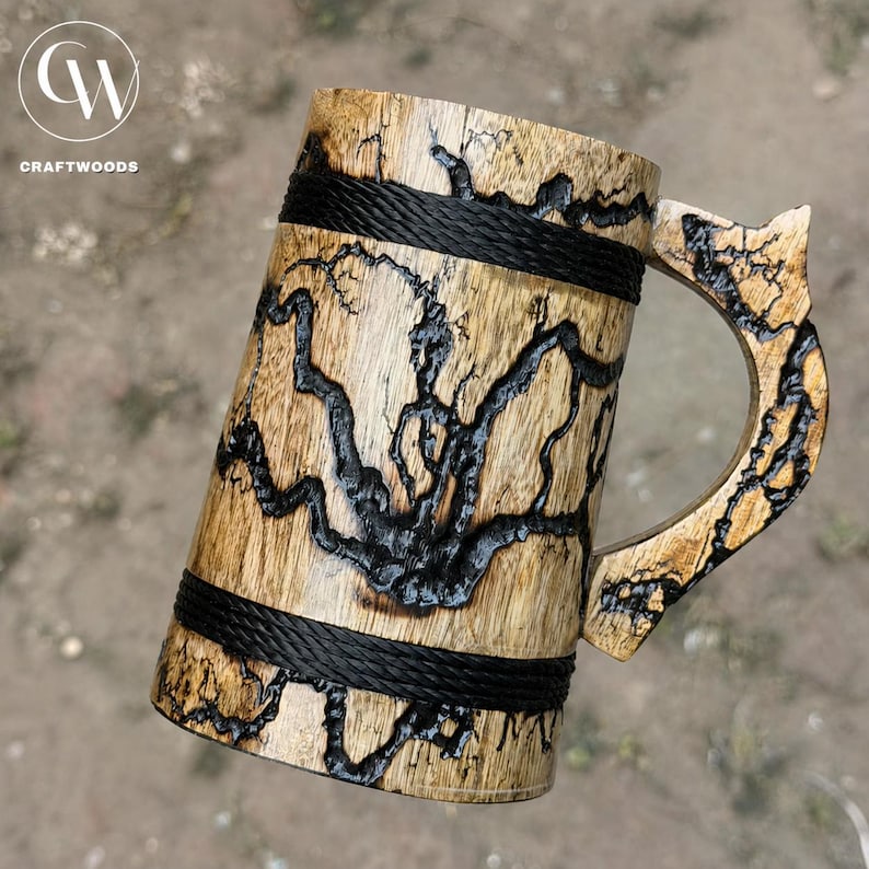 Pu&ograve; includere: Un boccale in legno caratterizzato da intricati motivi di bruciatura frattale nera sulla superficie. Il corpo cilindrico &egrave; accentuato da due fasce orizzontali in corda nera e include un manico in legno abbinato con un'estensione superiore a punta.