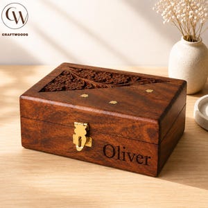 May include: A rectangular dark brown wooden box featuring a hinged lid with intricate floral carvings on one side and small brass flower inlays on the other. The front of the box has a gold-coloured metal latch and the engraved name Oliver.