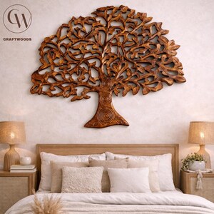May include: A detailed, carved wooden tree wall hanging in a warm brown hue. The tree features intricate leaf and branch patterns. The Craftwoods logo is visible. The artwork is displayed above a bed with neutral-toned bedding and bedside lamps.