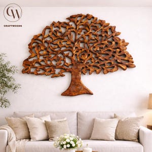 May include: A large, intricately carved wooden tree wall art piece. The tree is a warm brown colour, with detailed leaves and branches. The Craftwoods logo is in the upper left corner. The art is mounted on a white wall above a sofa with neutral-coloured cushions.
