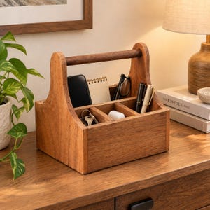 Op de afbeelding: Een middelbruine houten bureau-organizer met een centraal handvat en drie aparte vakken. De organizer bevat een zwarte smartphone, een notitieblok met spiraalbinding, een bril, twee pennen, een sleutelbos en een klein wit doosje.