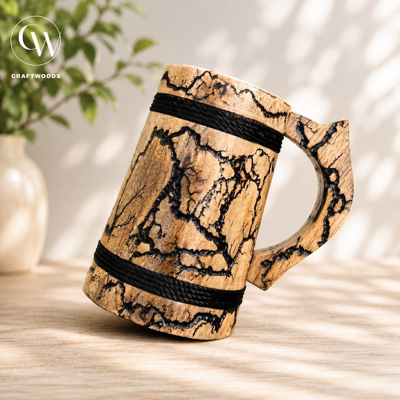 Pu&ograve; includere: Un boccale in legno caratterizzato da una finitura color legno naturale chiaro con motivi di bruciatura frattali scuri e ramificati sulla superficie. Il boccale &egrave; accentuato da due fasce orizzontali in corda nera avvolte attorno alla base e alla parte superiore.