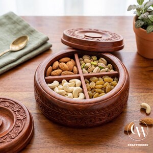 Op de afbeelding: Een ronde houten serveerschaal met vier compartimenten, gevuld met amandelen, pistachenoten, cashewnoten en rozijnen. De schaal heeft een decoratief gesneden ontwerp en een bijpassende deksel. Een gouden lepel en groene doek zijn op de achtergrond.