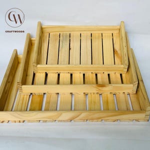 Puede incluir: Tres bandejas de madera de diferentes tamaños apiladas. Están hechas de madera clara con un diseño de listones en la parte inferior. El logotipo de Craftwoods está en la esquina superior izquierda.