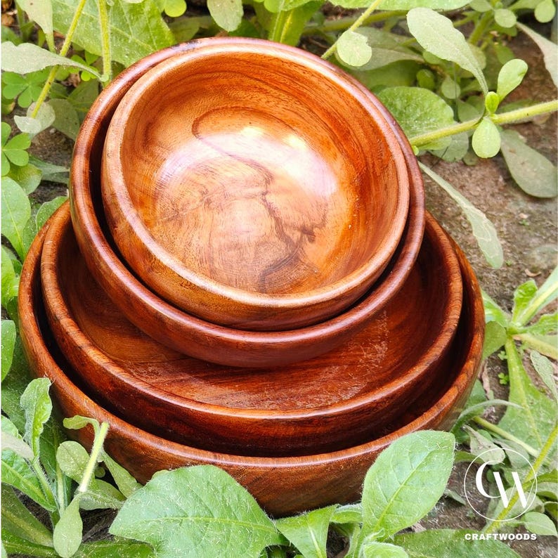 May include: A set of four wooden bowls nested together. The bowls are a warm brown color with visible wood grain. The bowls are of varying sizes, with the smallest bowl nested inside the others. The bowls are surrounded by green foliage.