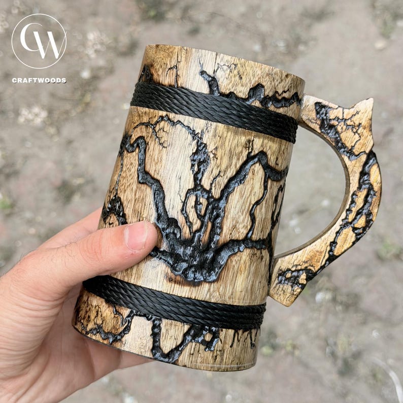 Pu&ograve; includere: Un boccale in legno con venature marrone chiaro naturale e intricati motivi di bruciatura frattale scuri sulla superficie. Il boccale presenta due fasce orizzontali in corda nera attorno al corpo e un manico in legno abbinato con dettagli.