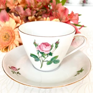 Könnte beinhalten: Weißes Tee-Service mit einer Tasse und Untertasse, verziert mit einem rosa Rosenmuster und goldenen Rändern. Die Tasse zeigt eine detaillierte Rosenillustration, die Untertasse ein kleineres Rosenmotiv. Das Set ist wahrscheinlich aus Porzellan.