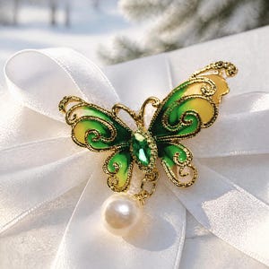 May include: A gold-toned butterfly brooch featuring green and yellow enamel wings with filigree detailing. The center of the butterfly is adorned with a small green gemstone, and a single white faux pearl hangs from the bottom of the brooch.
