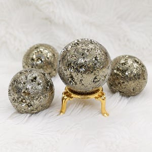 Könnte beinhalten: Vier kugelförmige Pyritsteine mit einer metallischen, strukturierten und kristallinen Oberfläche. Eine Pyritkugel ist auf einem kleinen, dreibeinigen goldfarbenen Metallständer erhöht, während die anderen drei Kugeln darum angeordnet sind.