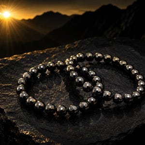 Könnte beinhalten: Zwei kreisförmige Armbänder aus dunklen, metallischen, facettierten, kugelförmigen Perlen. Die Perlen haben eine reflektierende, polierte Oberfläche, die das Licht einfängt und einen starken Kontrast zur dunklen, strukturierten Steinoberfläche bildet.