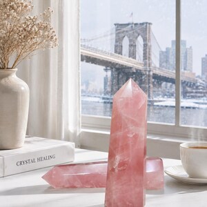 Könnte beinhalten: Zwei polierte Rosenquarz-Kristalltürme mit sechseckiger Basis und spitzer Oberseite. Ein Turm steht vertikal, während der andere horizontal neben einem weißen Buch mit dem Titel Crystal Healing, einer Keramikvase und einer Teetasse liegt.