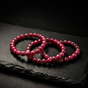 Könnte beinhalten: Zwei kreisförmige Armbänder aus facettierten, tiefroten Edelsteinperlen. Die Perlen haben eine reflektierende, mehrfach facettierte Oberfläche, die das Licht einfängt und ein strukturiertes Erscheinungsbild auf den runden, gleichmäßigen Formen erzeugt.
