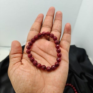 Könnte beinhalten: Ein kreisförmiges Perlenarmband bestehend aus zahlreichen kleinen, runden, facettierten tiefroten Edelsteinen. Die Perlen sind von einheitlicher Größe und auf ein elastisches Band gefädelt, was eine durchgehende Schlaufe aus dunkelroten Steinen bildet.