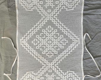 May include: White crochet apron with a detailed openwork design. The apron features a rectangular shape with a decorative pattern and long ties for securing around the waist. The apron is made of a white yarn.