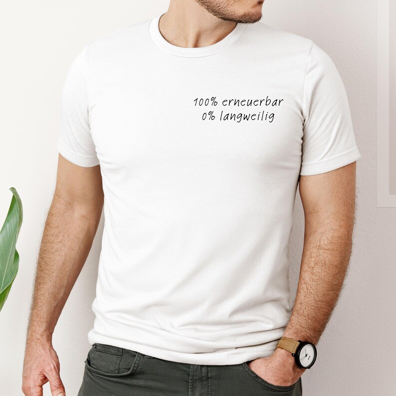 Puede incluir: Camiseta blanca con el texto "100% erneuerbar 0% langweilig" en escritura negra. La camiseta es de manga corta y parece estar hecha de un material suave. La persona lleva un reloj con esfera negra y correa beige.