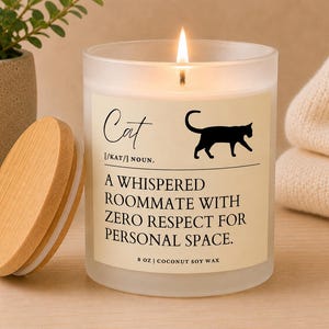 May include: A frosted glass candle with a lit wick and a wooden lid. The label features a black cat silhouette and the text: Cat [/kat/] noun. A whispered roommate with zero respect for personal space. 8 oz | coconut soy wax.