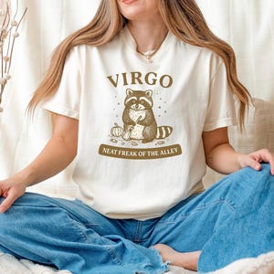 May include: Cream-colored t-shirt featuring a raccoon illustration with the text "VIRGO" above and "NEAT FREAK OF THE ALLEY" below. The raccoon is holding a pumpkin and a broom. The shirt is made of a soft material.