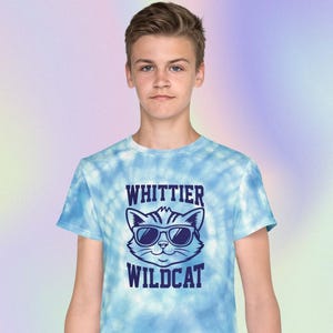May include: A light blue tie-dye t-shirt featuring a graphic of a cat wearing sunglasses with the text "Whittier Wildcat" in navy blue. The shirt is made of soft material and is suitable for casual wear.