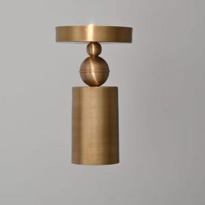 May include: A brass-colored metal candle holder featuring a cylindrical base, a central spherical element with a smaller sphere above it, and a flat circular top platform. The object has a smooth, matte finish and a vertical, stacked geometric design.