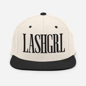 Puede incluir: Gorra de béisbol color crema con visera plana negra. La palabra "LASHGRL" está escrita en negro y en negrita en la parte delantera. La parte superior de la gorra tiene un botón negro.