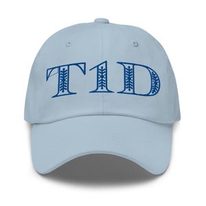 May include: A light blue baseball cap with the embroidered text "T1D" in a dark blue, decorative font. The cap has a curved brim and a button on top. The hat is made of a soft material.