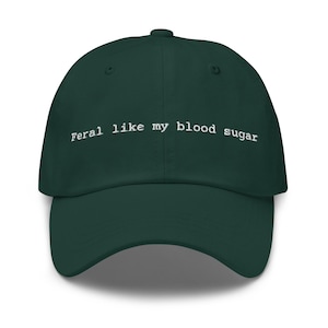 May include: A dark green baseball cap with the text "Feral like my blood sugar" in white. The cap has a curved brim and a button on top. The text is centered on the front of the cap.