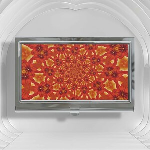 May include: A rectangular metal business card case featuring a polished silver finish and a central inset panel displaying a vibrant, repeating kaleidoscopic pattern of orange and red floral shapes radiating from the center of the design.