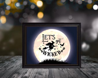 May include: A framed Halloween-themed print featuring a full moon with a witch on a broomstick and the words "Let's get Wicked." The black frame sits on a wooden surface, with a blurred background of bokeh lights.