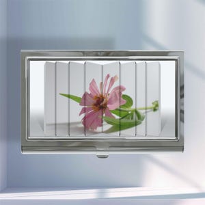 May include: A rectangular silver metal business card case featuring a glossy finish. The front surface displays a segmented, accordion-style print of a single pink zinnia flower with green leaves, creating a layered visual effect across the metal.