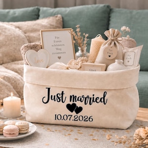 Può includere: Un cesto regalo in tessuto beige con le parole "Just married" e la data 10.07.2026 in corsivo nero. Il cesto contiene vari articoli a tema matrimonio, tra cui candele, una decorazione a forma di cuore e piccoli sacchetti regalo. Ci sono dei macarons davanti.