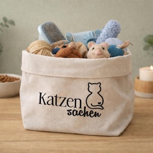 Könnte beinhalten: Ein beiger Aufbewahrungskorb aus Segeltuch, gefüllt mit Katzenspielzeug, einer Bürste und einer Schnurkugel. Der Korb trägt die Aufschrift "Katzen Sachen" und eine schwarze Katzensilhouette.